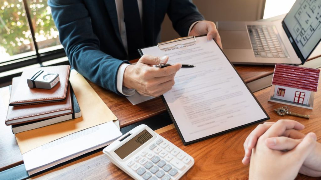 Un conseiller bancaire évalue un contrat d'achat immobilier en présence d'un client, avec des documents, une maison miniature et une calculatrice sur la table.